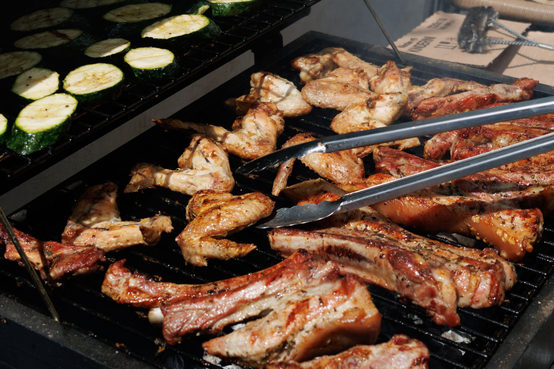 Tongs turning ribs and chops on a grill beside courgette slices